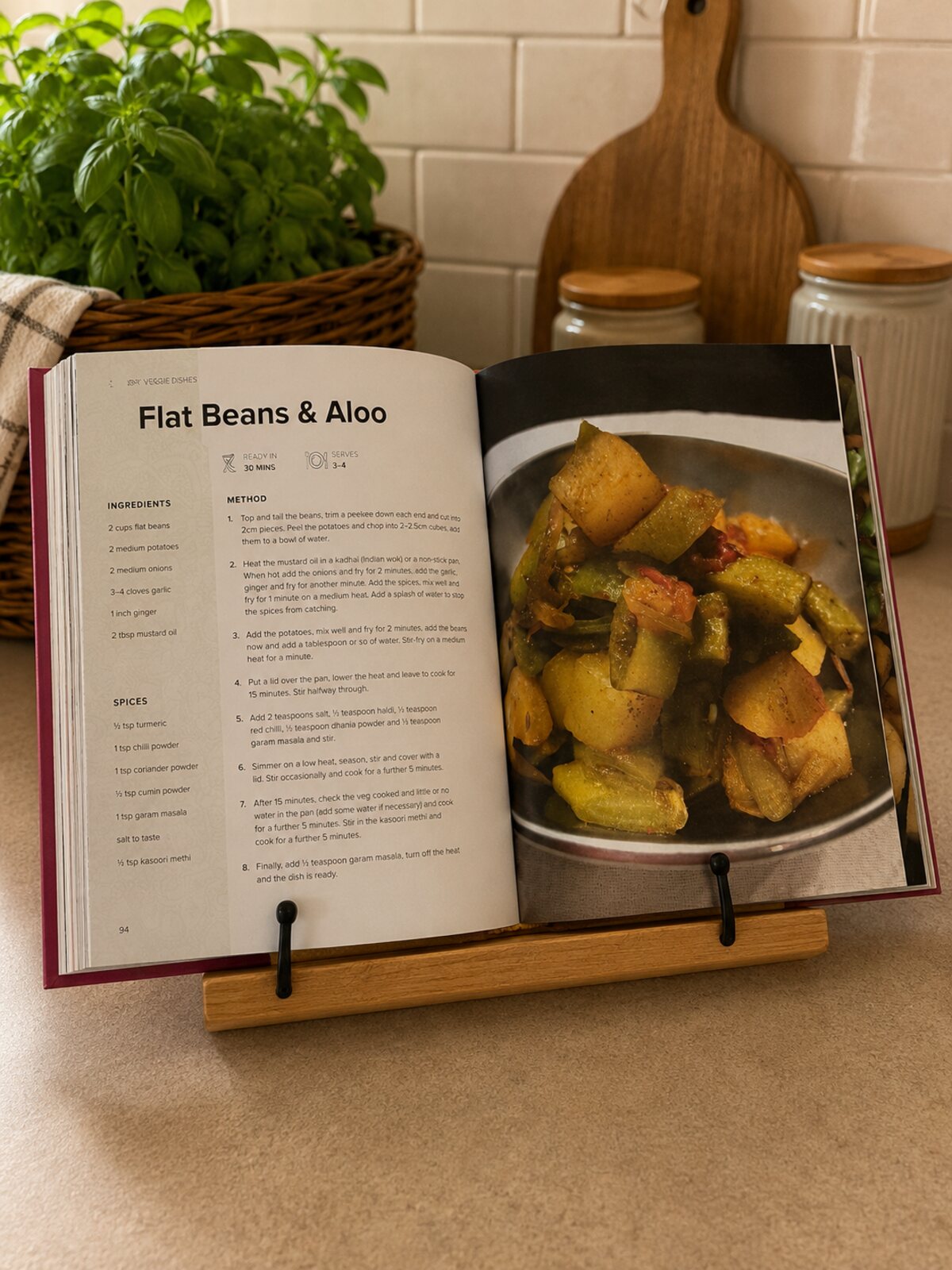 The book open on a wooden stand at the Flat Beans and Aloo recipe, with ingredients, spices and a photo of the finished dish
